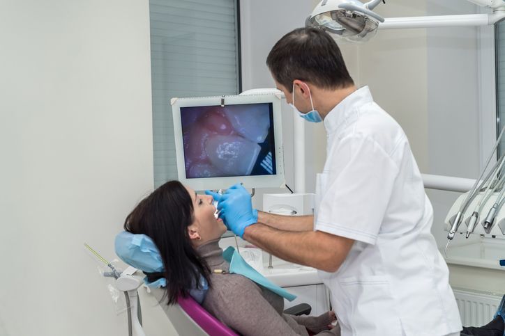 Dentist examining a patient’s mouth in a bright dental clinic, with a monitor showing X-ray images