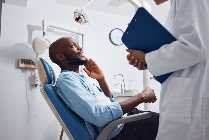 Doctor speaking to patient in a clinic, holding a blue clipboard while seated patient listens in a recliner.