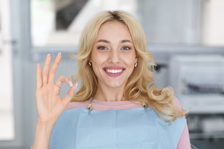 Smiling blonde woman in a blue apron making an OK gesture in a bright kitchen