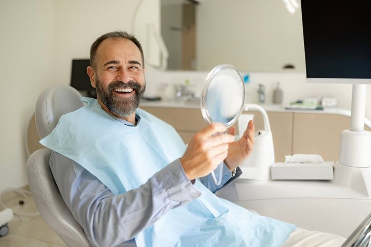 Man reclining in a dental chair, smiling while holding a mirror in a bright clinic