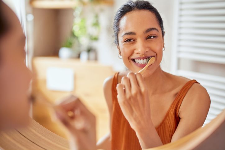 Woman smiling while applying lipstick in a mirror, wearing an orange top in a bright room