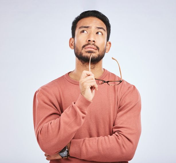 Man in a rust sweater thinking with glasses held to his chin against a plain light background