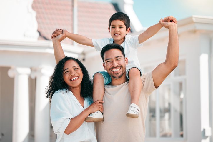 Smiling family posing outside, with a child on each parent’s shoulders in bright daylight