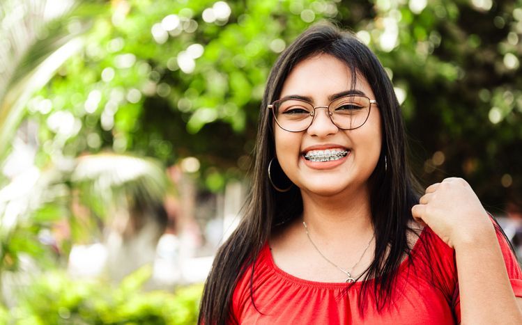 Smiling person in a red top and glasses outdoors with blurred greenery in the background