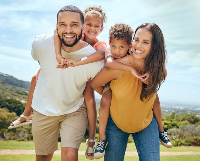 Smiling family of four outdoors on a grassy hill, with parents carrying two children on their backs.
