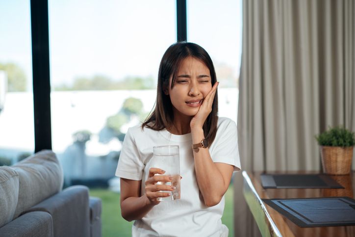 Woman holding her cheek and a drink in a bright living room, appearing uncomfortable.
