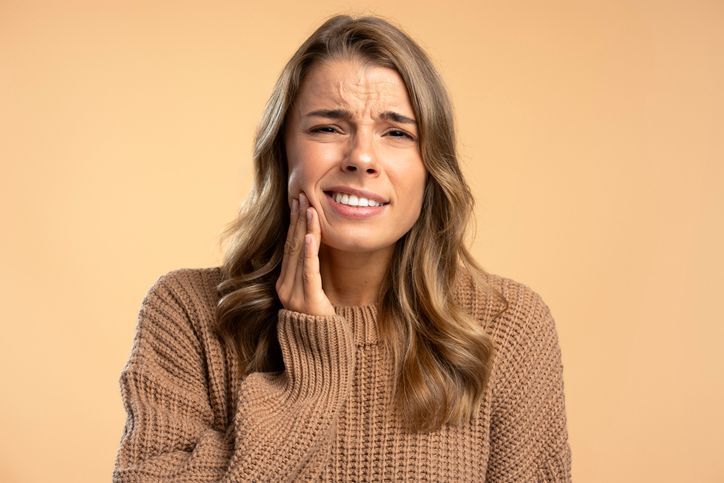 Person in a brown sweater holding their cheek and grimacing against a beige background
