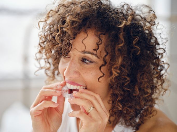 Woman smiling while putting in clear aligners in a bright bathroom