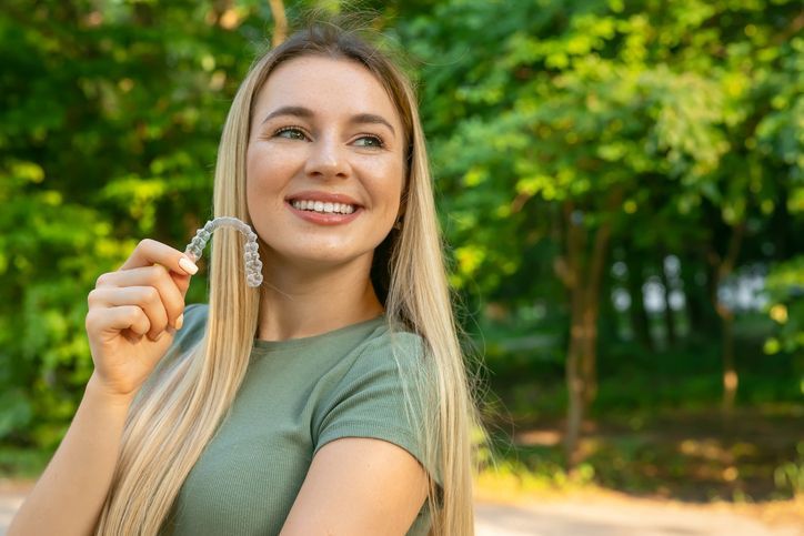 Smiling woman holding clear dental aligners outdoors in a green park