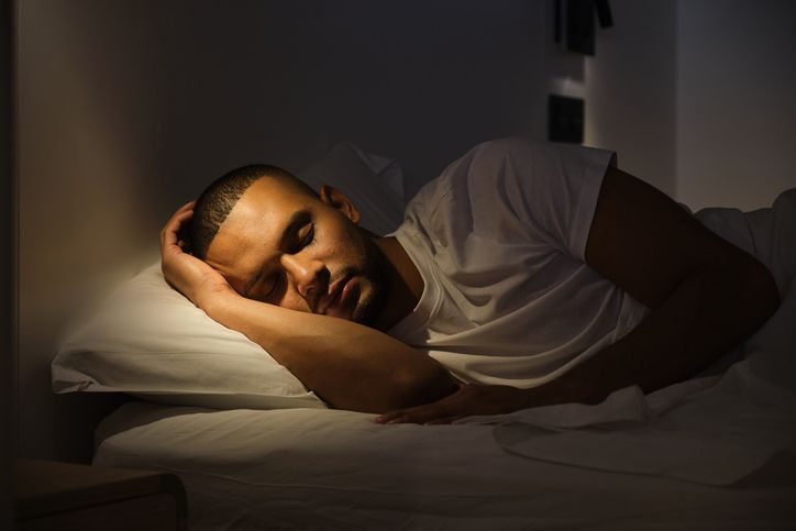 Man lying on a bed in dim light, resting with one arm under his head.