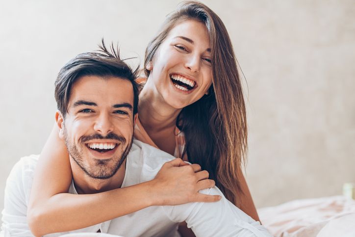 Two smiling people embracing on a bed in a bright, cozy room