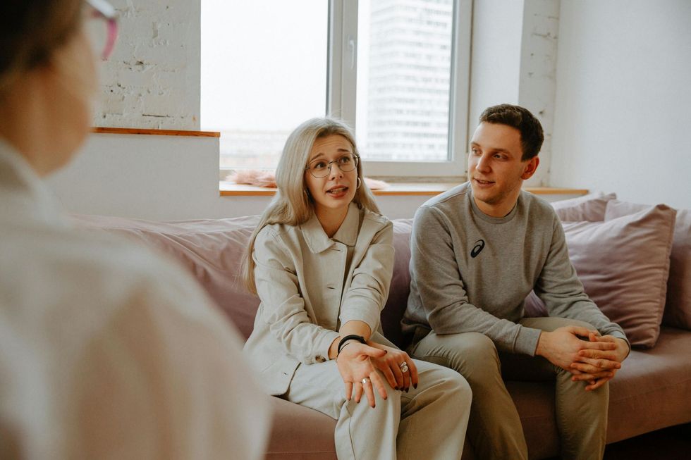 Couple talking to therapist on a couch; woman in glasses, man, and therapist partially visible.