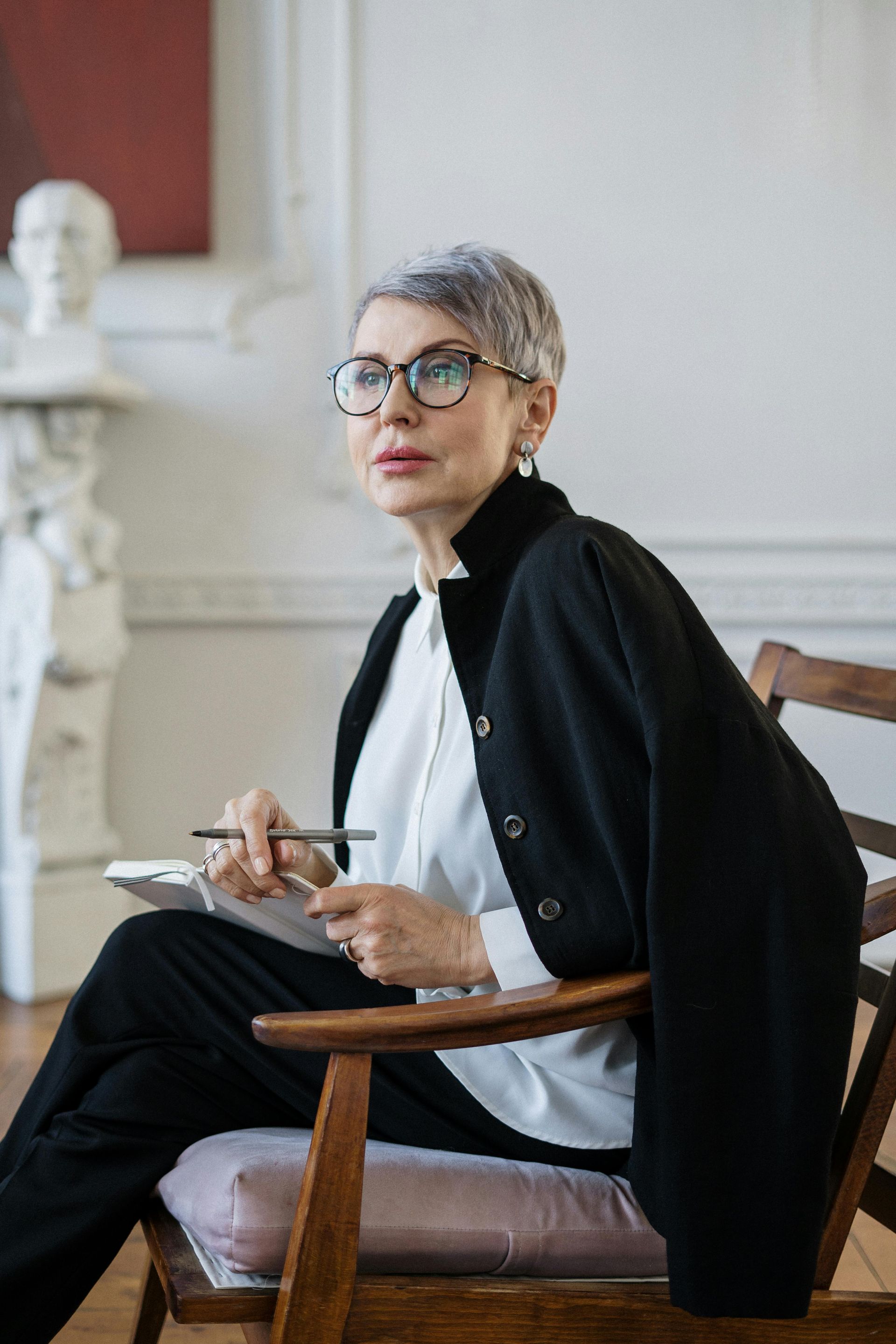 Woman with gray hair and glasses sitting in a chair, holding pen and notebook, looking thoughtful.