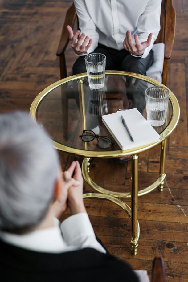 Therapist and client in discussion, sitting across a small table with water, notepad, and glasses.
