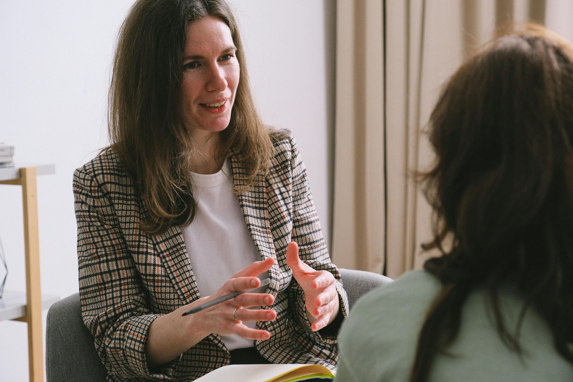Woman in blazer gestures, talking to another person in an office setting.