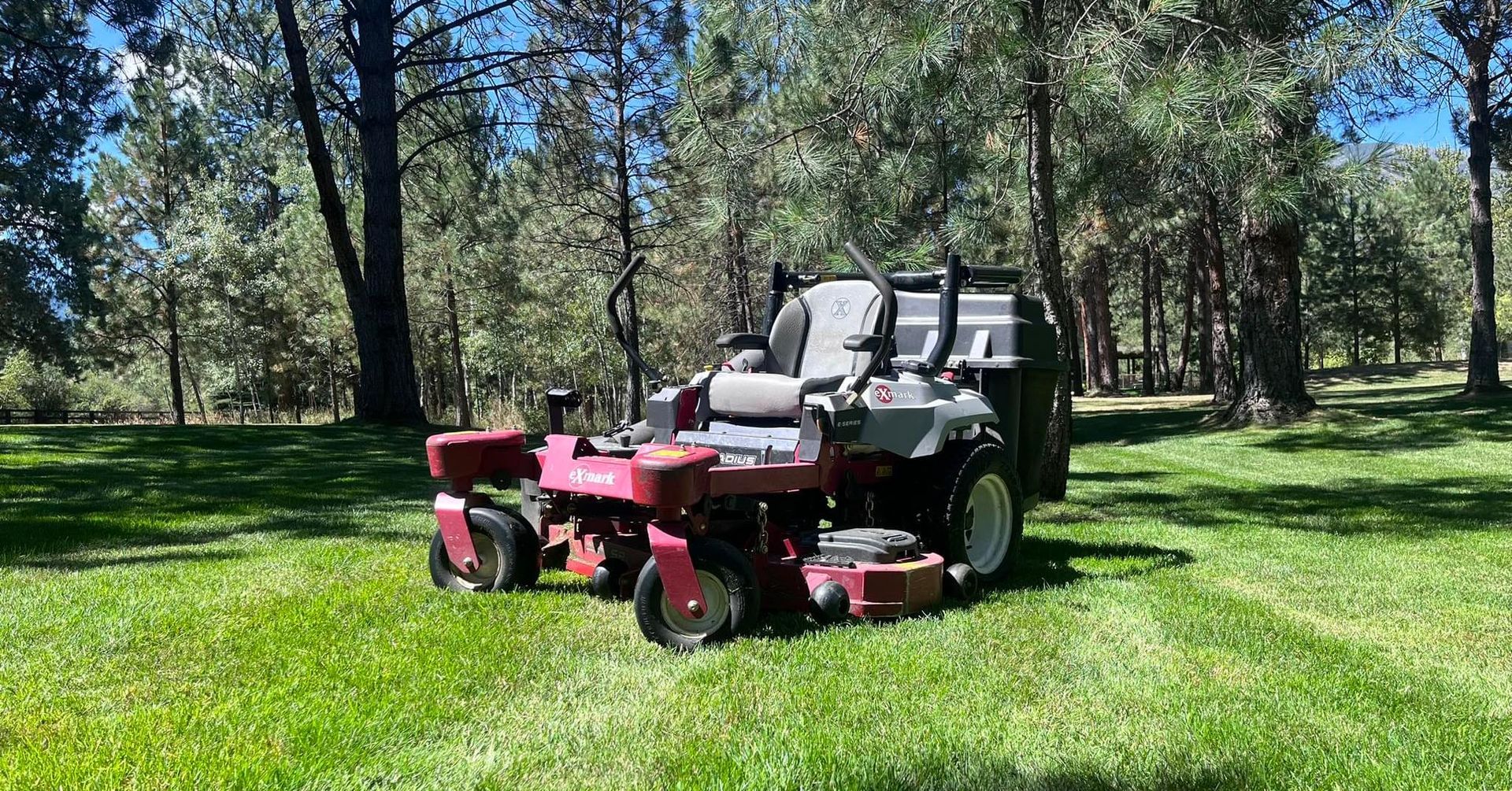 A red zero-turn mower sits on a grassy lawn surrounded by trees on a sunny day.