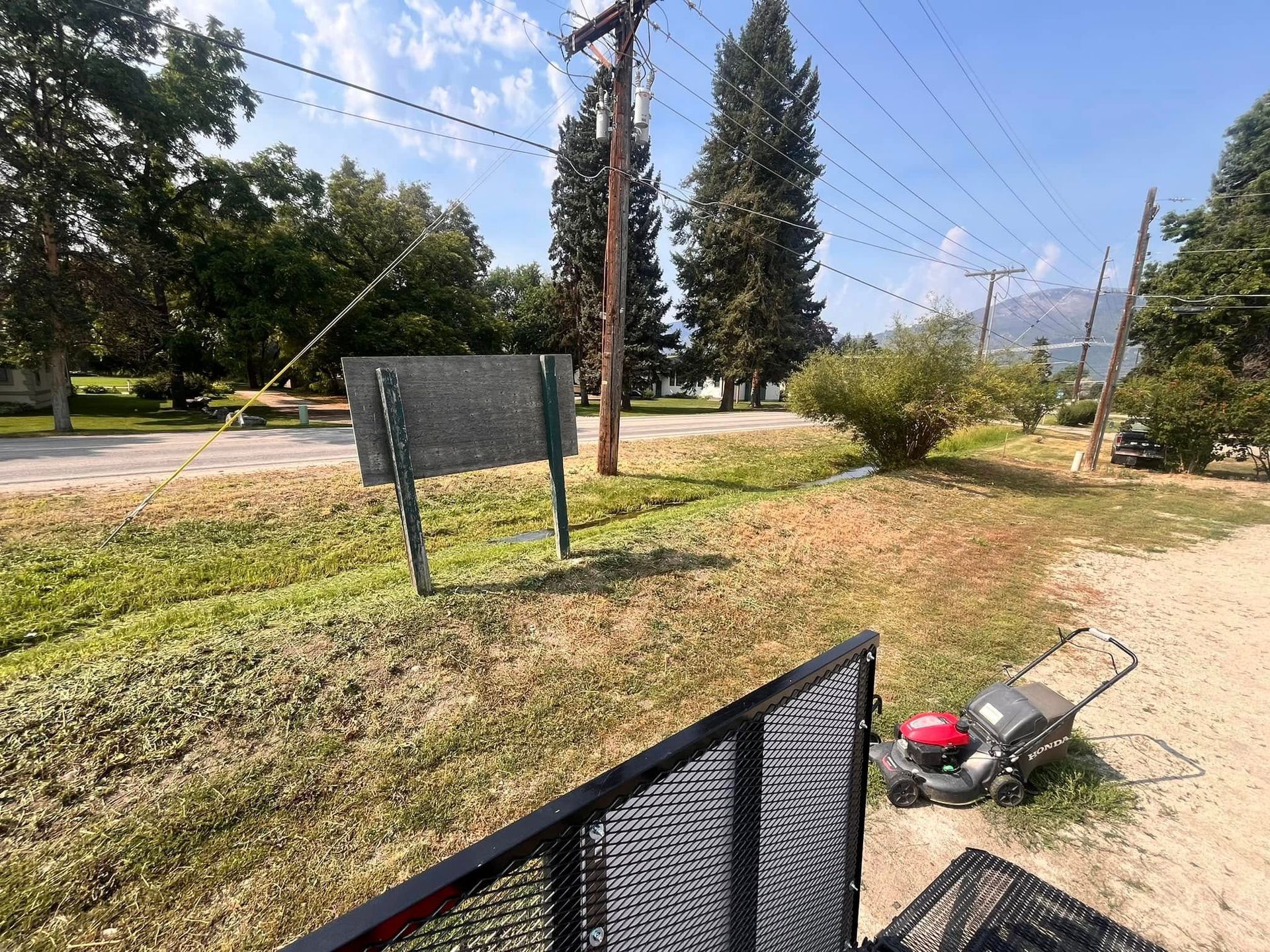A lawnmower sits on the grass near a metal trailer gate, facing a roadside area with a blank sign and utility poles.
