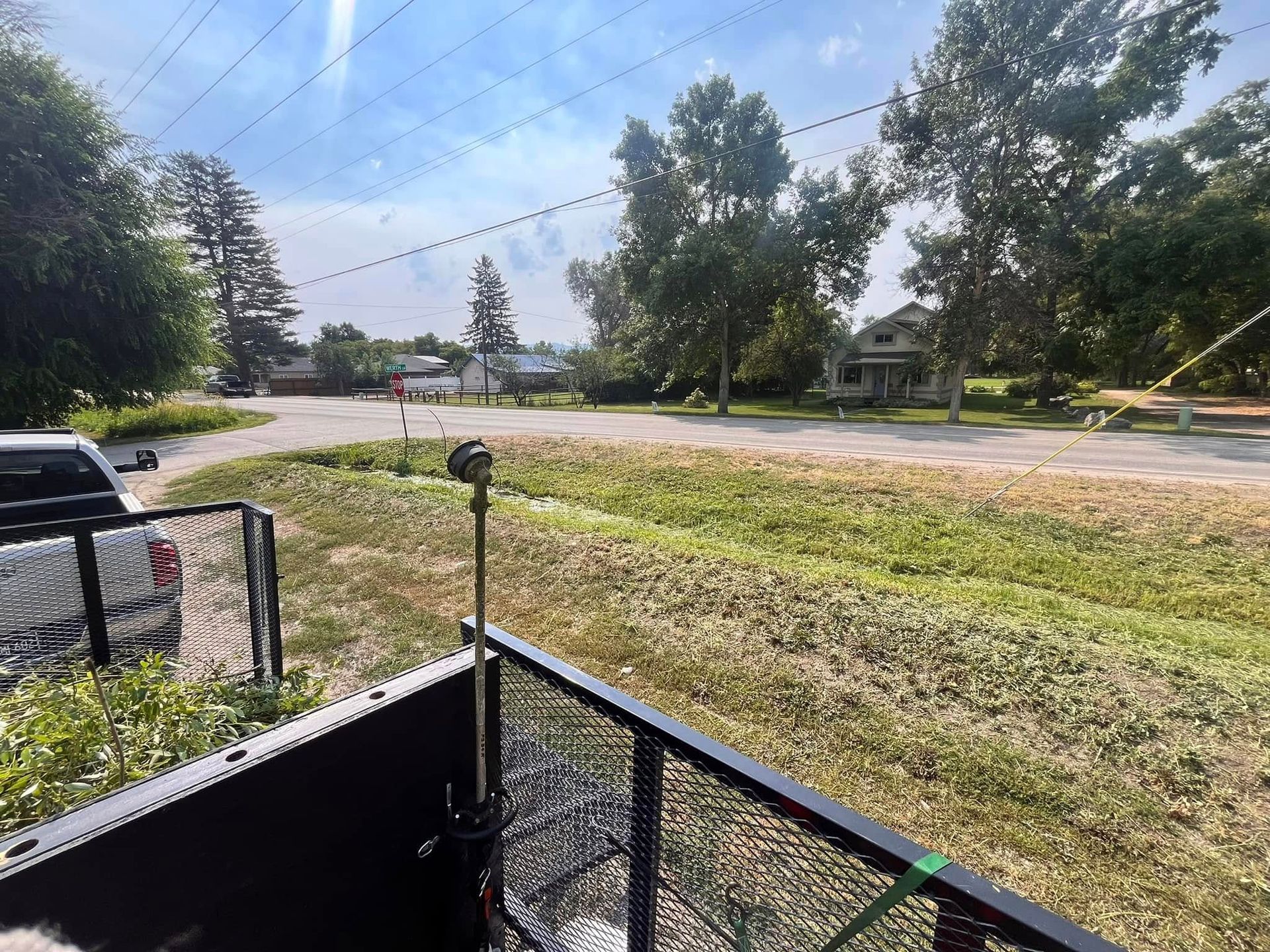 View from the back of a truck, showing a metal pole, a grassy field, a quiet road, trees, and a house in the distance.
