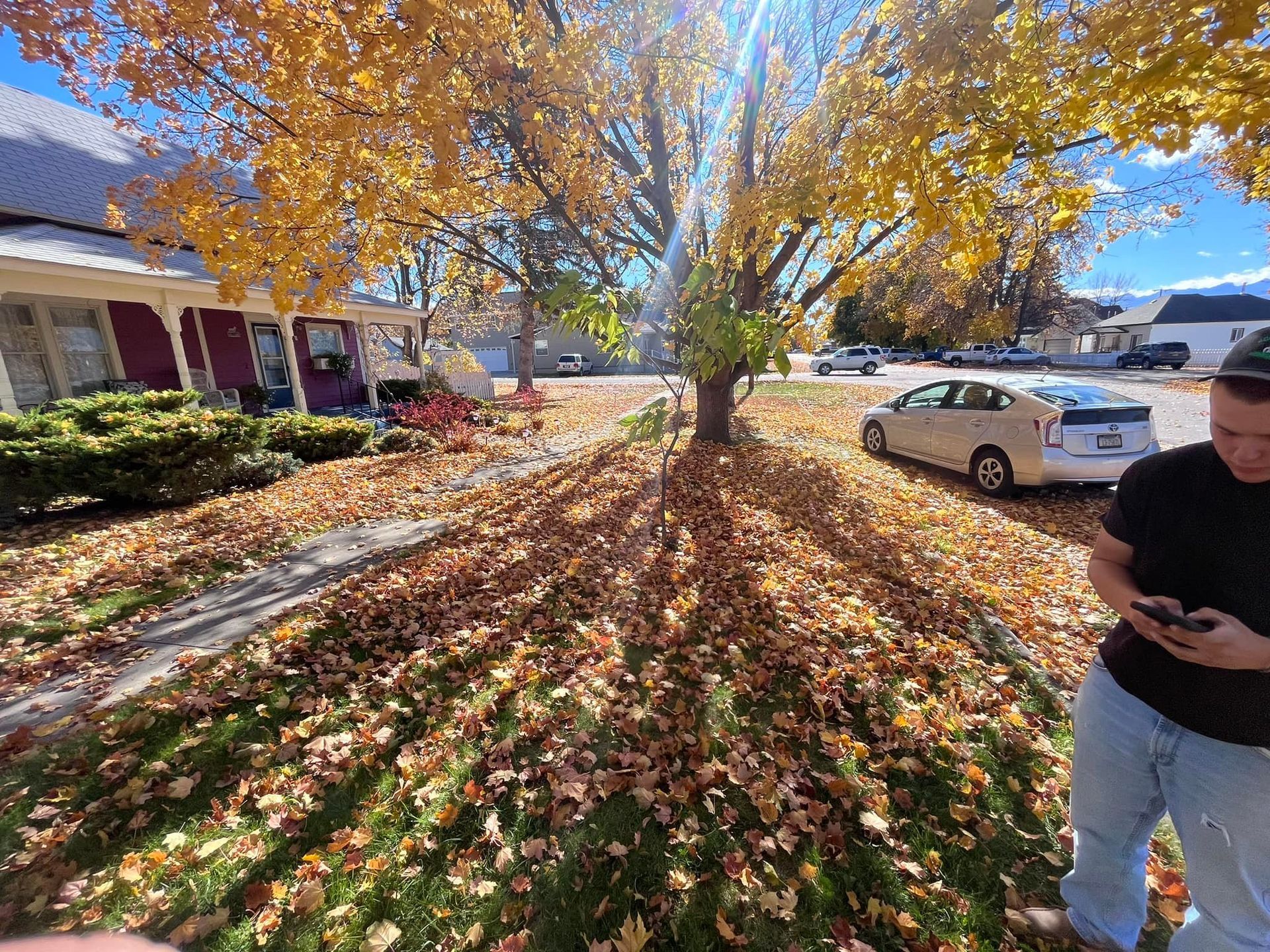 A person stands in a yard covered in autumn leaves in front of a house, with a parked car on the street nearby.