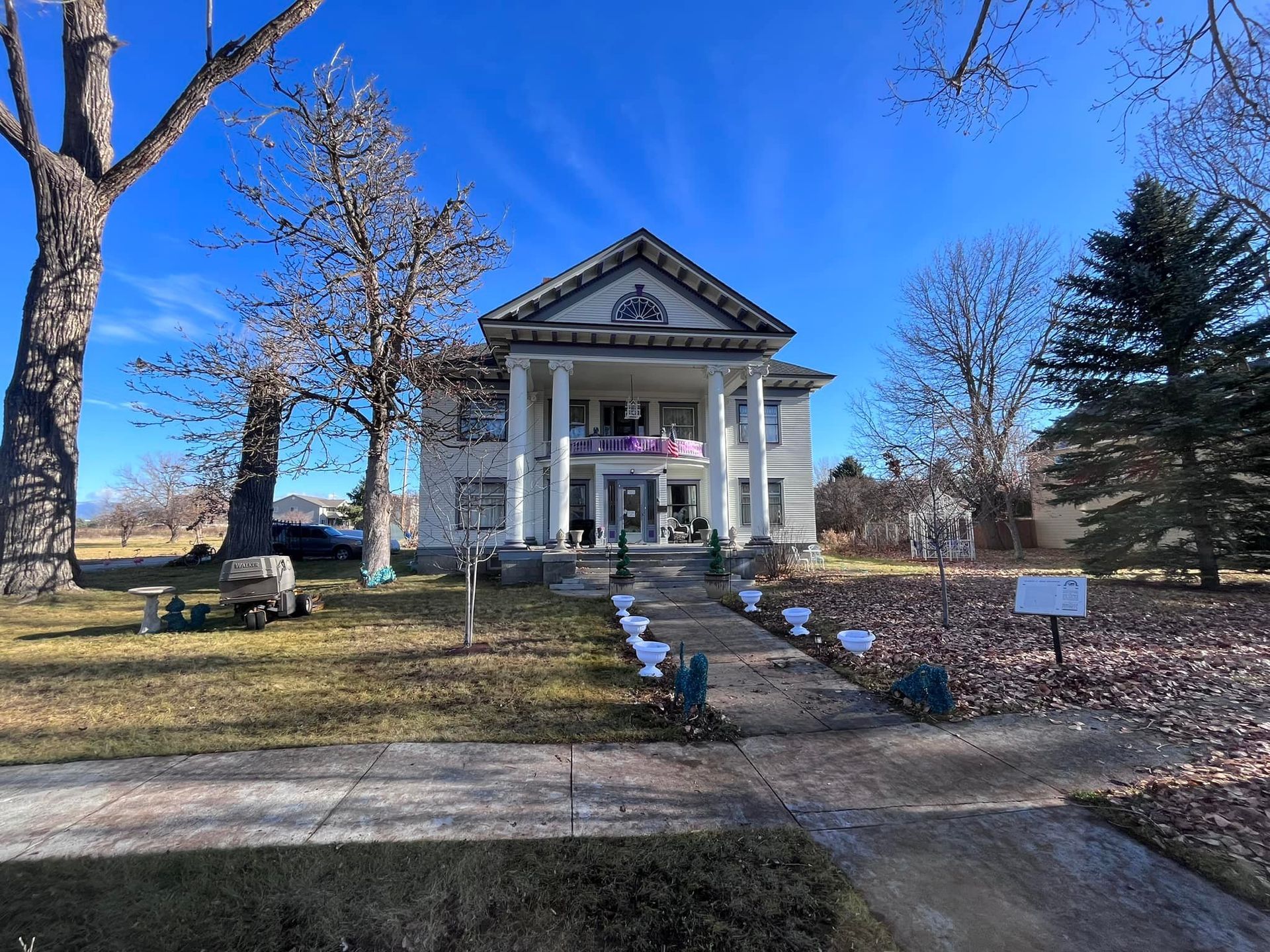 A two-story white house with a columned front porch and a triangular gable roof, set on a lawn with trees on a sunny day.