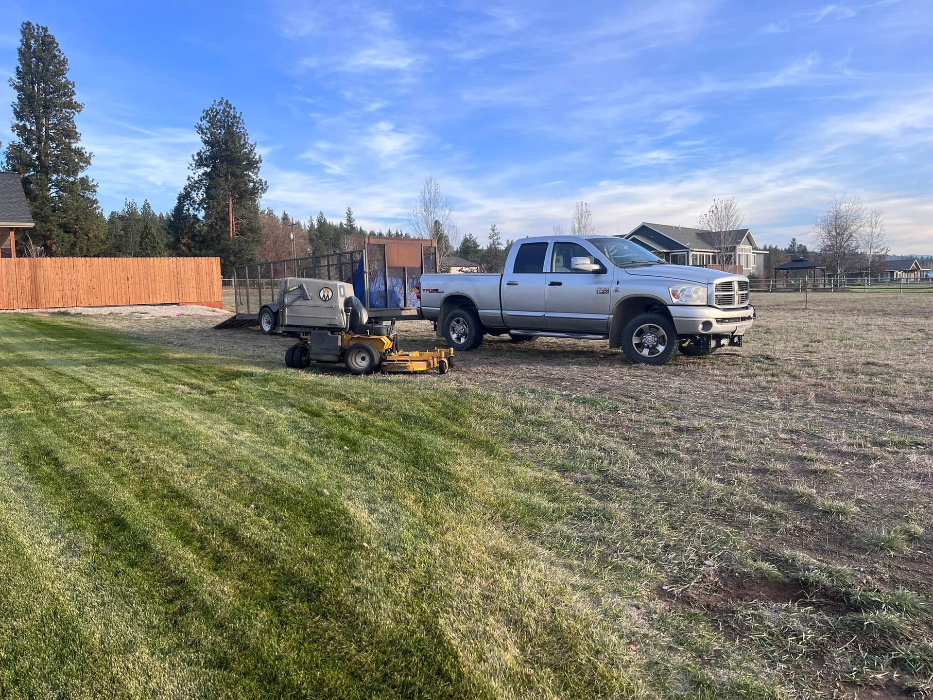 A silver pickup truck with a trailer and yellow lawn mower parked on a field next to a freshly cut green lawn.