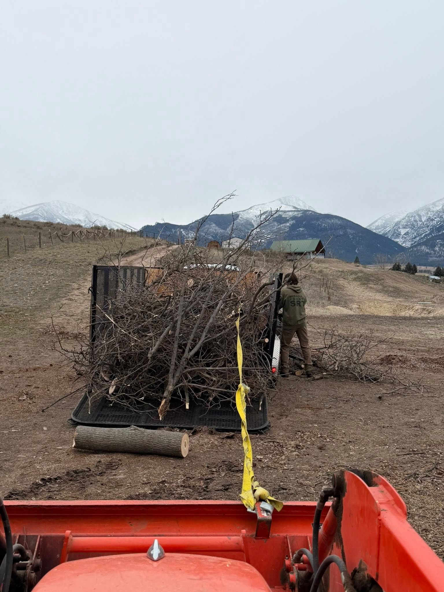 A person in camouflage gear pulls a trailer filled with brush behind a red tractor in a rural landscape with mountains.
