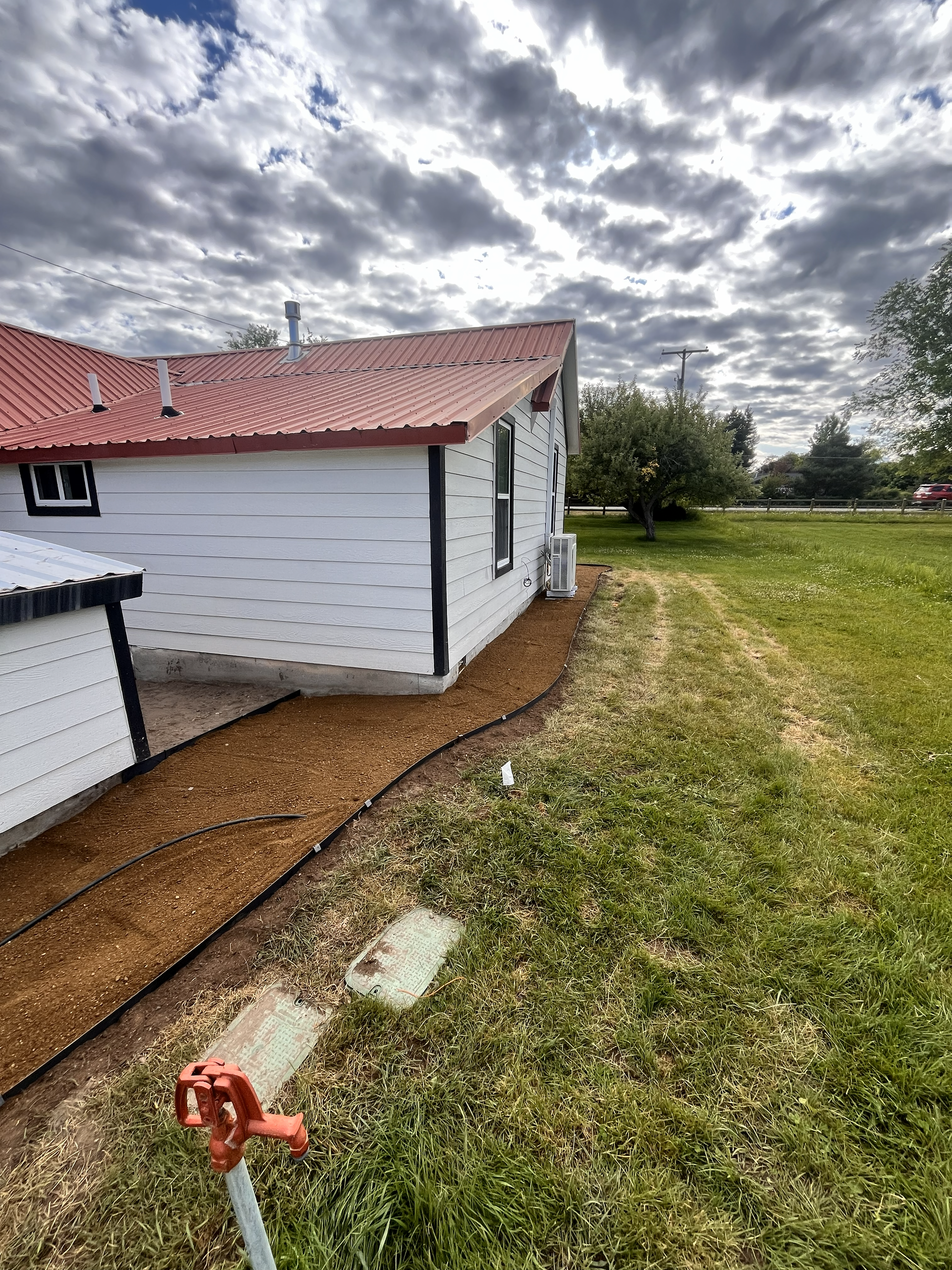 A white house with a red metal roof and newly laid wood chip landscaping beside a grassy lawn under a cloudy sky.