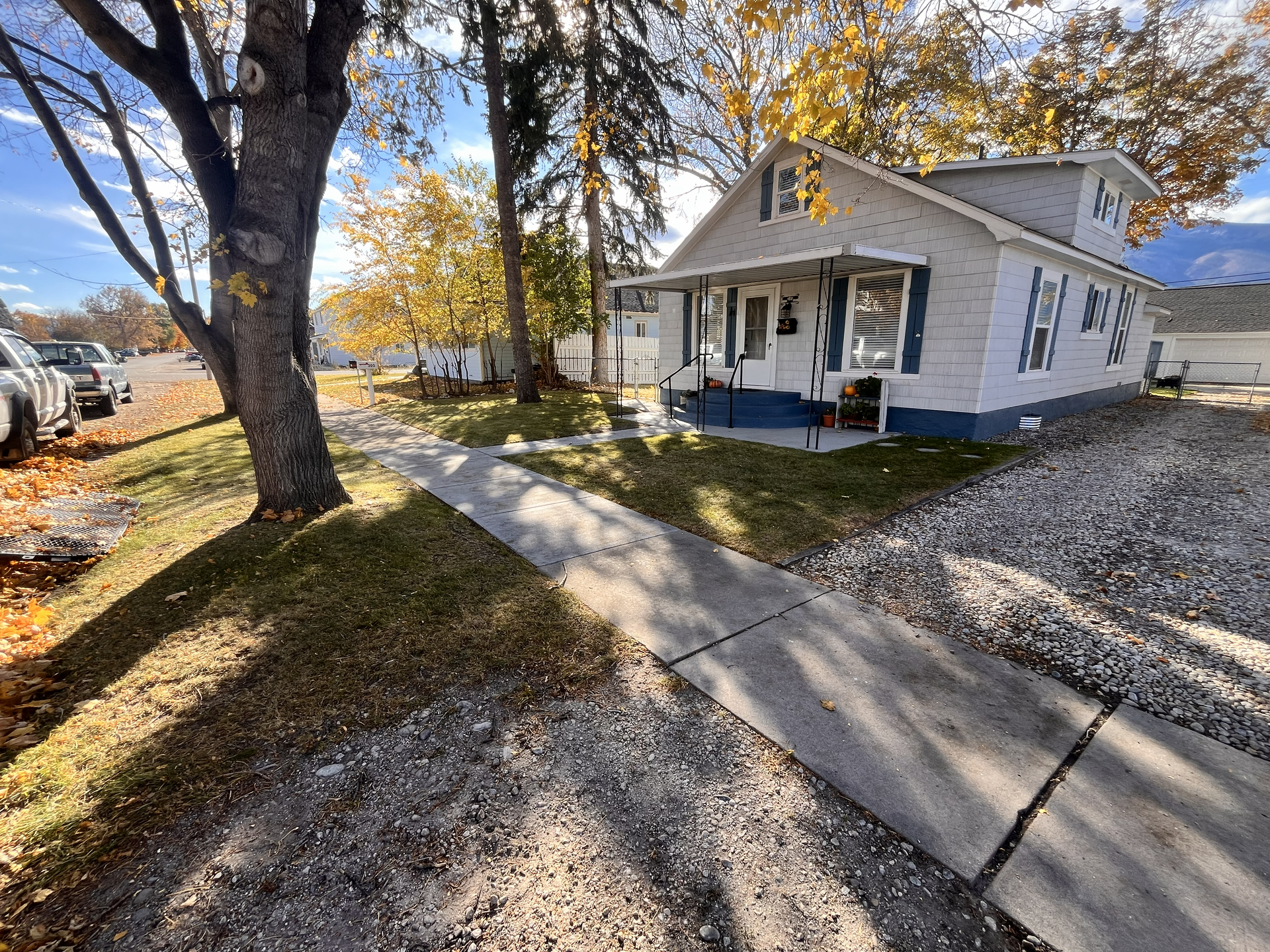 A white one-story house with a blue foundation sits behind a sidewalk on a sunny, autumn day with golden trees.