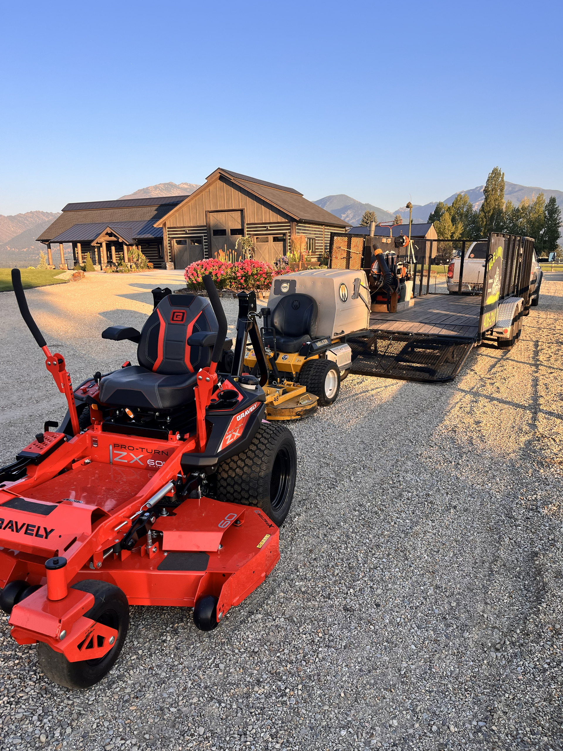A bright red zero-turn mower sits on a gravel lot in front of a trailer loaded with outdoor power equipment and buildings.