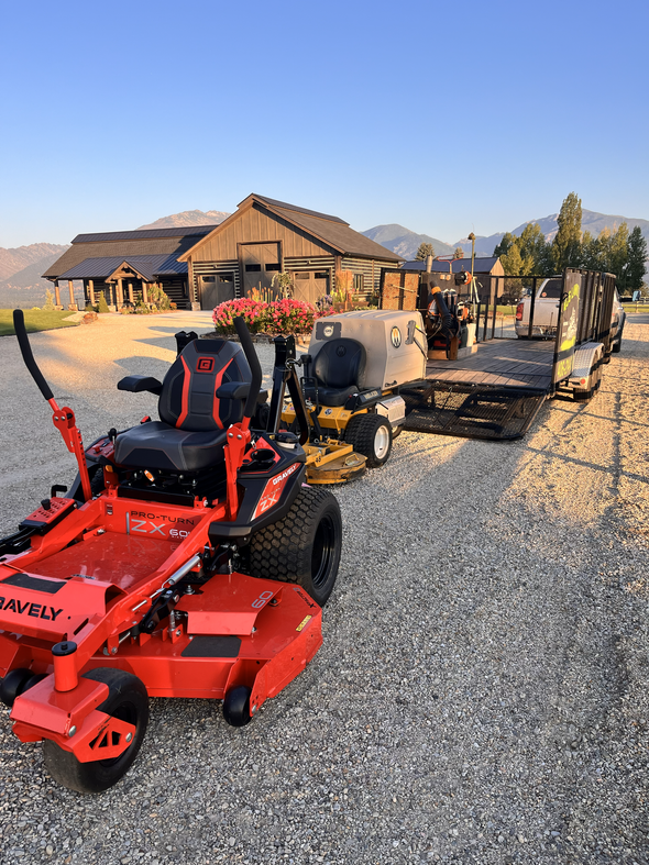A bright orange Gravely zero-turn mower sits on a gravel driveway in front of a flatbed trailer and rustic buildings.