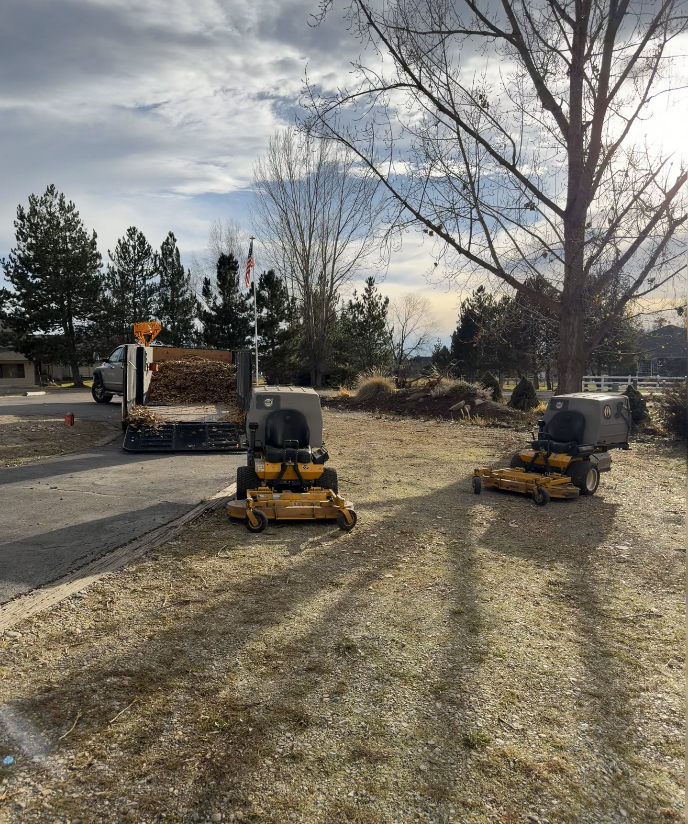 Two yellow zero-turn mowers parked on a dirt lot with a truck trailer loaded with yard debris in the background.