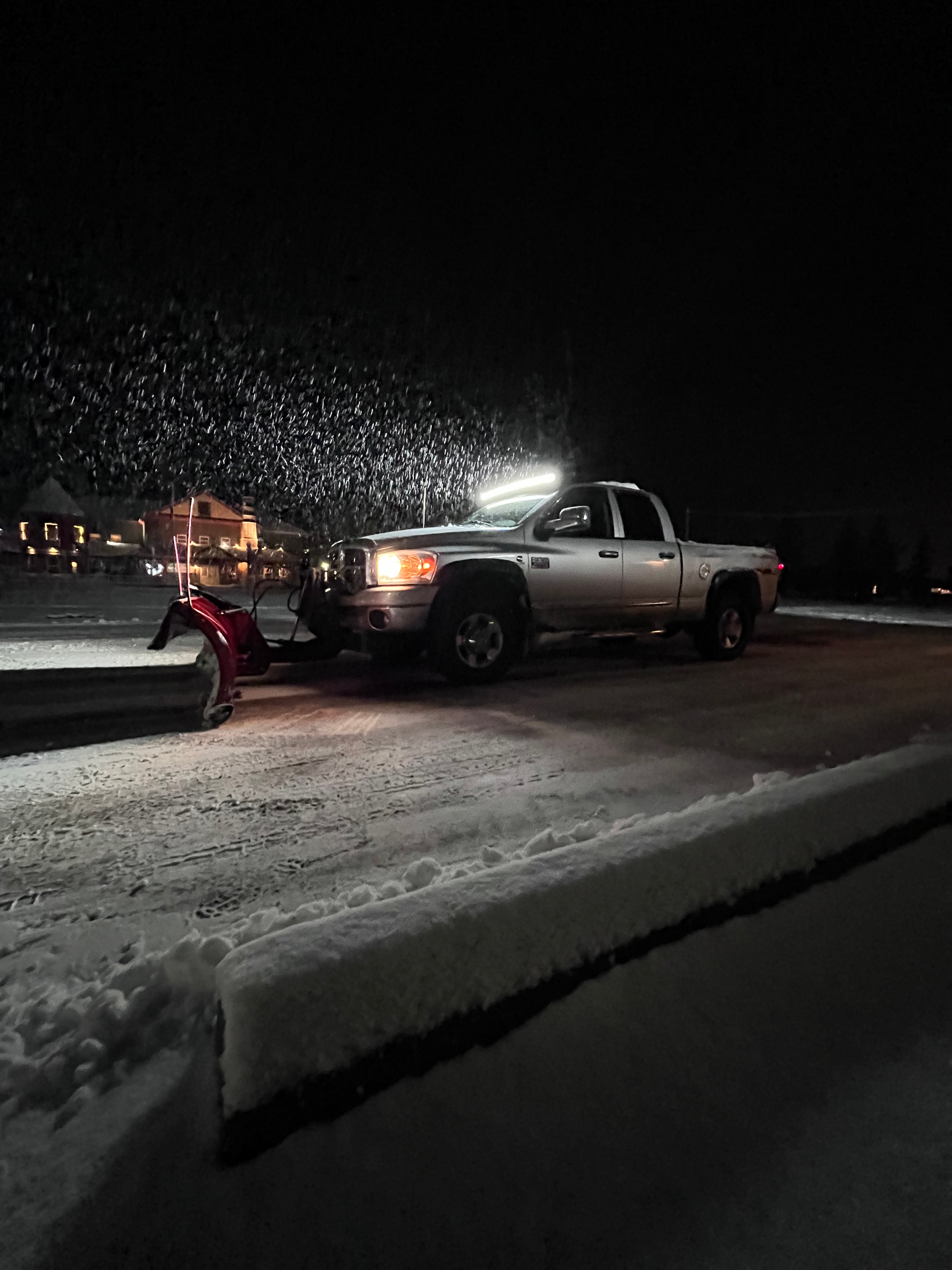 A silver pickup truck with a red snowplow attached to the front parked on a snowy road at night.