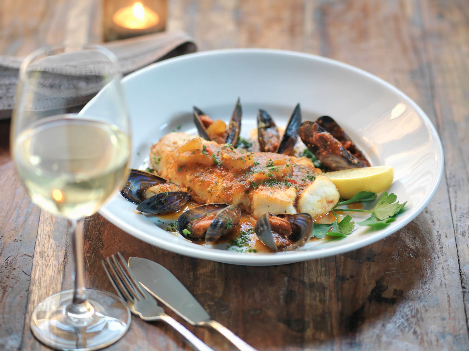 Pesce e cozze in salsa su un piatto bianco con vino bianco e posate su un tavolo di legno.