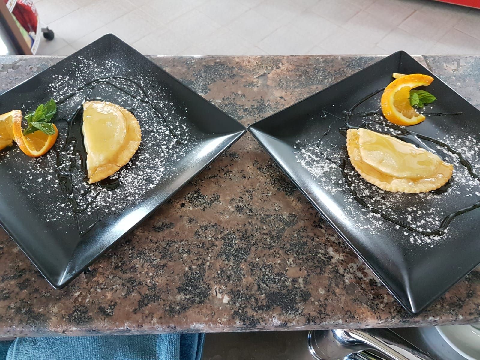 Due dessert su piatti quadrati neri con fette d'arancia e menta, spolverati con zucchero a velo, su un bancone.