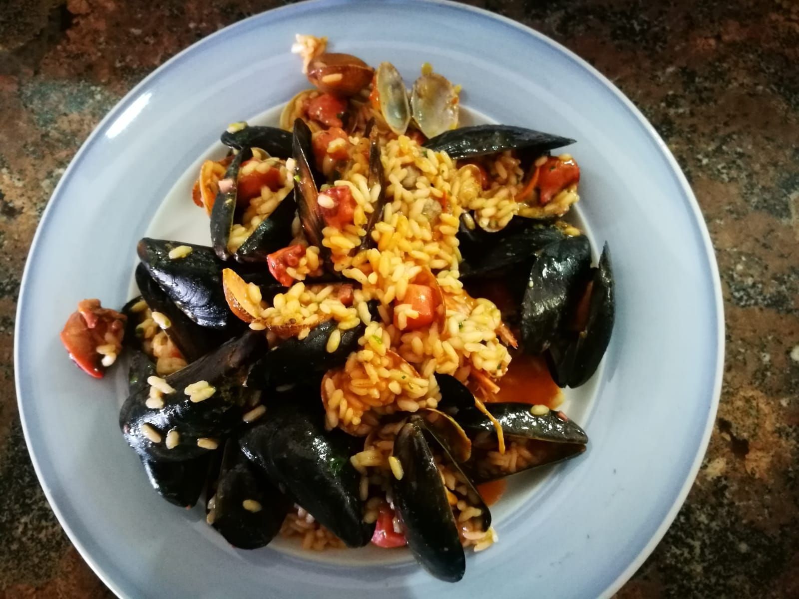 Piatto di riso e cozze su un piatto azzurro. Le cozze nere circondano il riso ricoperto di pomodori.