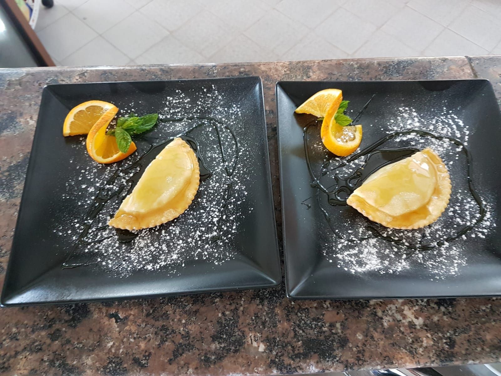 Due piatti quadrati neri, ciascuno con un dessert, fette d'arancia e zucchero a velo.