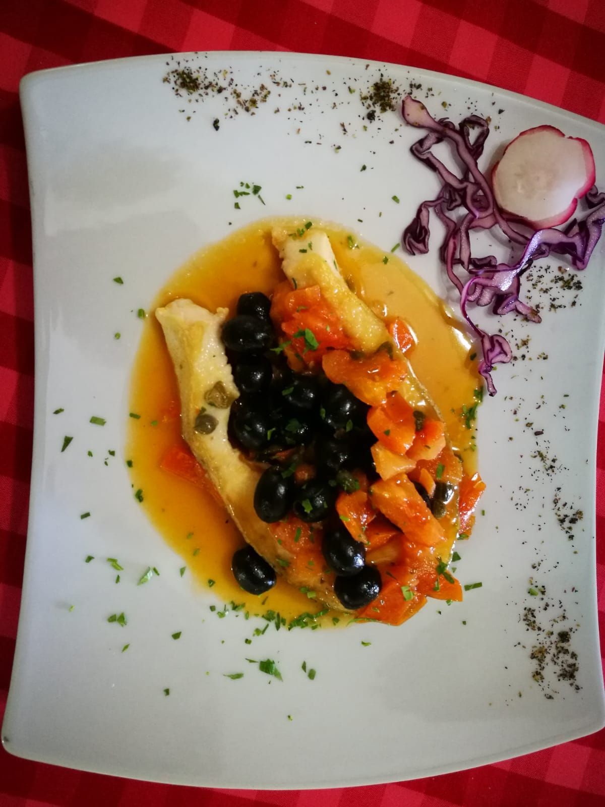 Filetto di pesce con salsa, olive e pomodori su un piatto bianco, guarnito con prezzemolo e cavolo rosso.