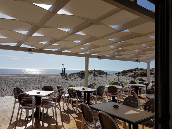 Bar all'aperto sulla spiaggia con tavoli, sedie e tenda da sole; sullo sfondo l'oceano e il cielo limpido.