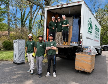 Five people in green shirts pose by a moving truck, with boxes and equipment on a driveway.