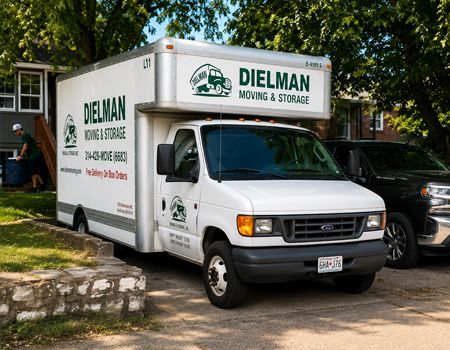 White Dielman moving truck parked in a driveway beside a black SUV, with trees and a house in the background