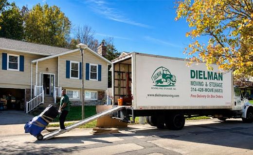 Mover unloading furniture from a truck in front of a beige house on a sunny day
