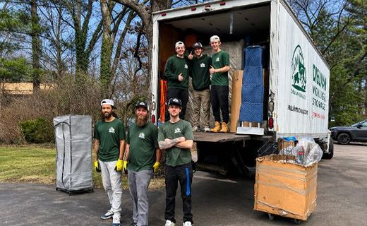 Five people unloading boxes from a moving truck on a tree-lined street.