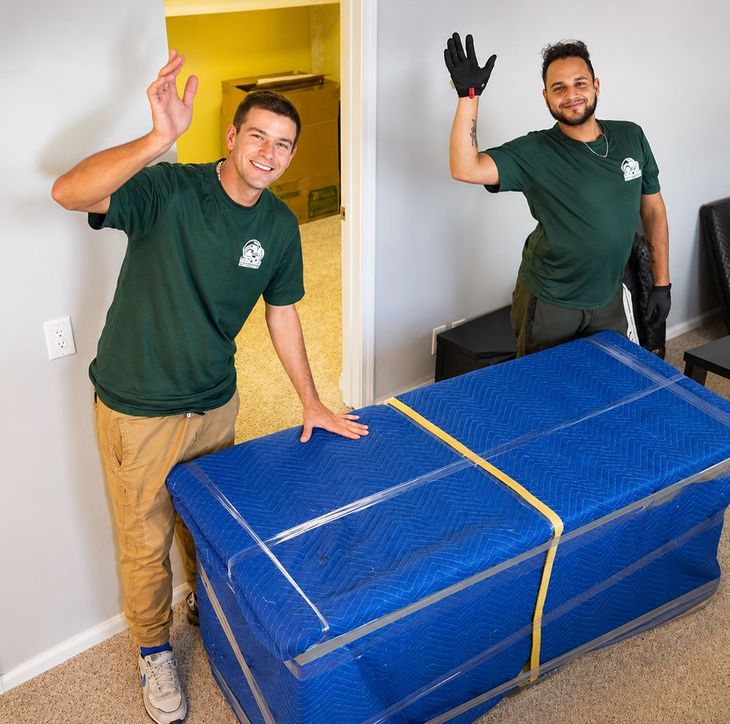 Two movers wave beside a large blue-wrapped item in a home interior.