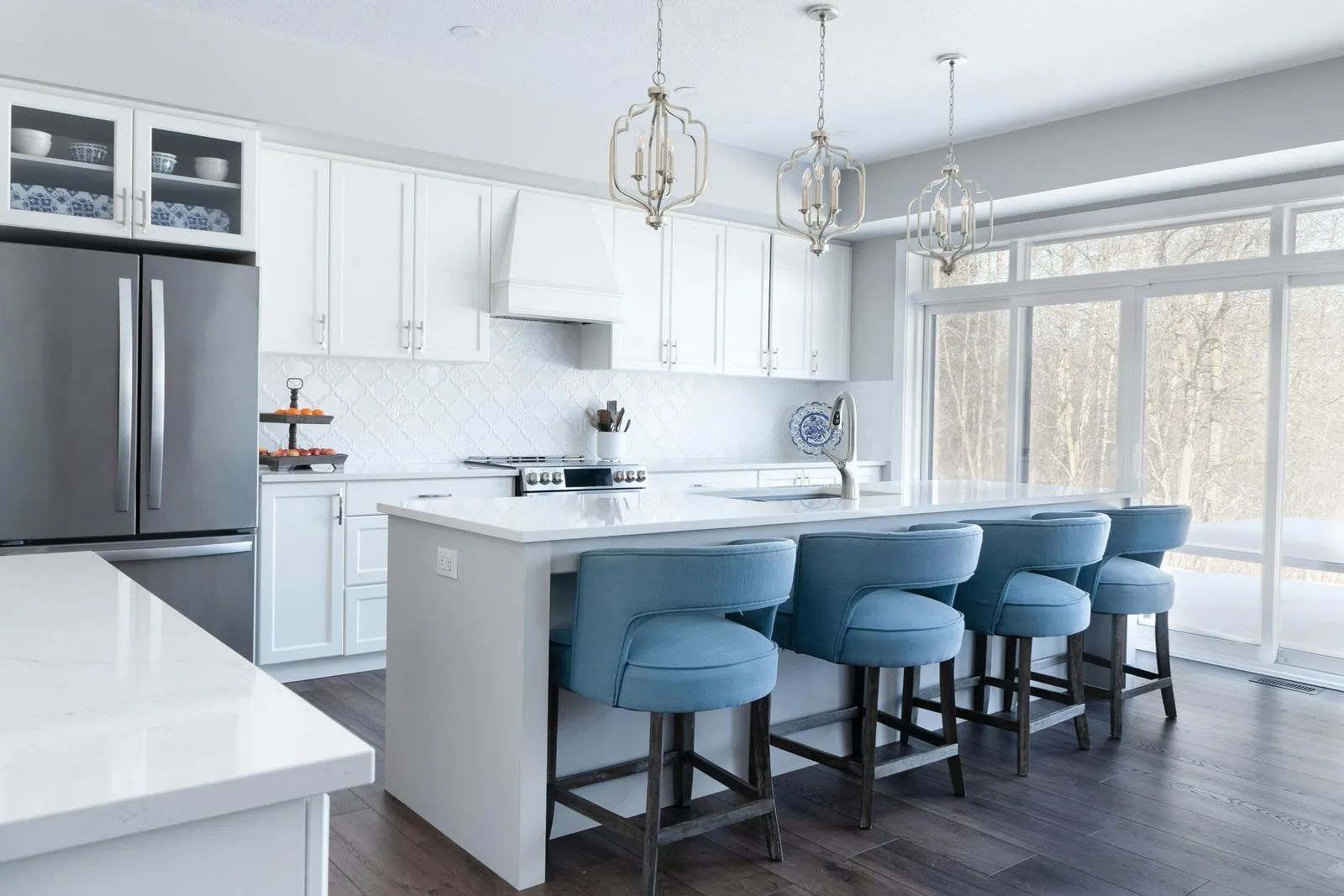 Bright modern kitchen with white cabinets, gray island, blue stools, and large windows.