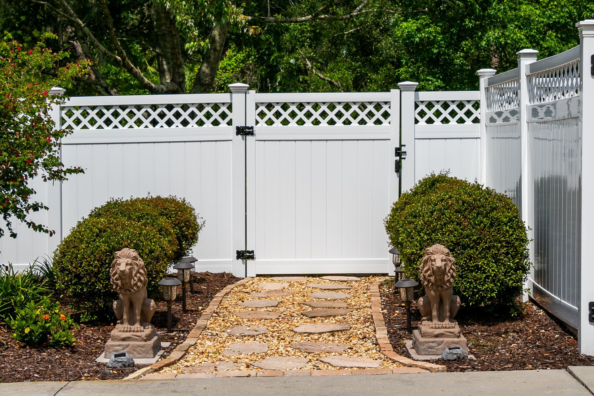 A white vinyl fence with a gate and a stone path flanked by two lion statues and rounded shrubs.