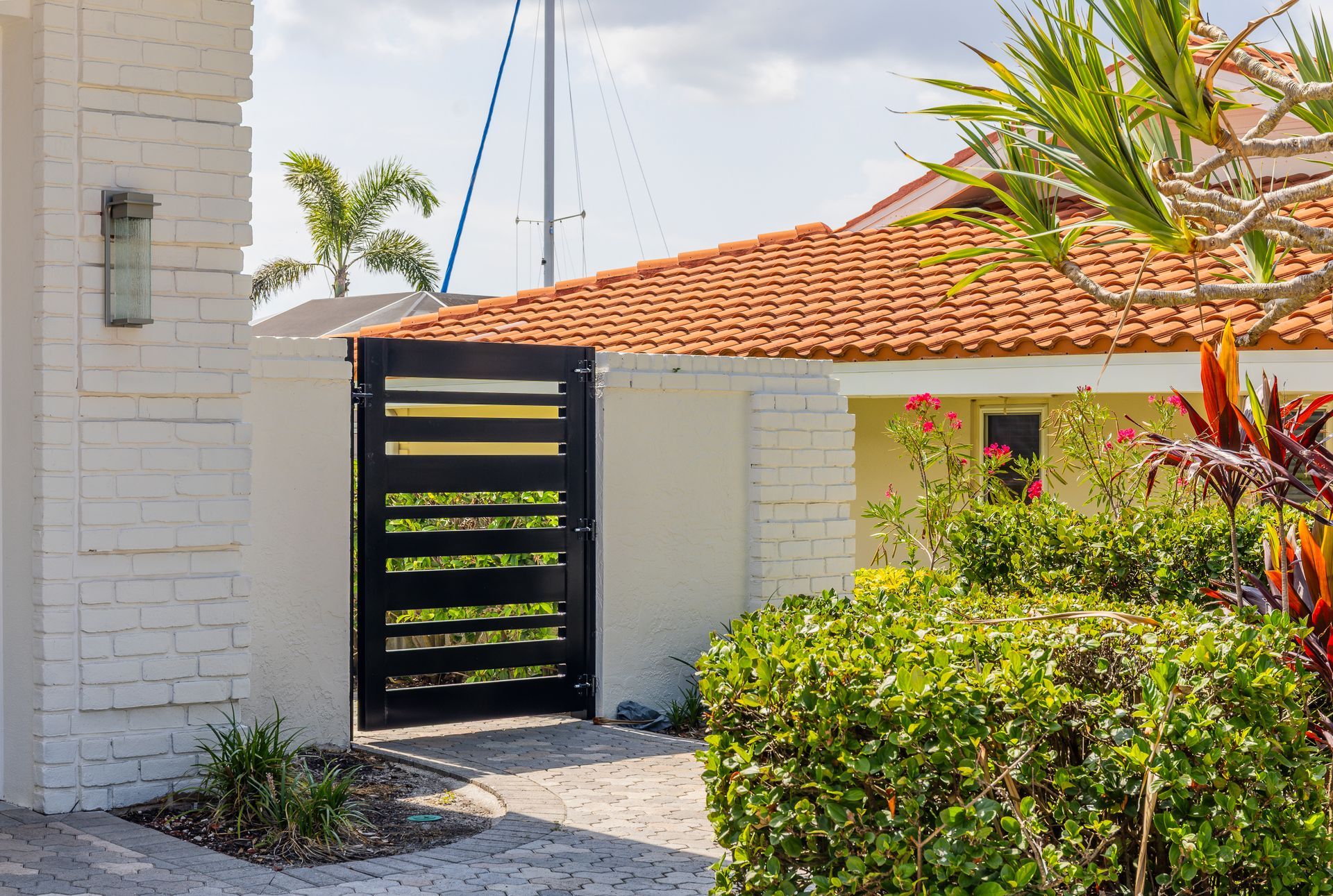 A black horizontal slat gate sits in a white painted brick wall next to a house with a terracotta tiled roof and bushes.