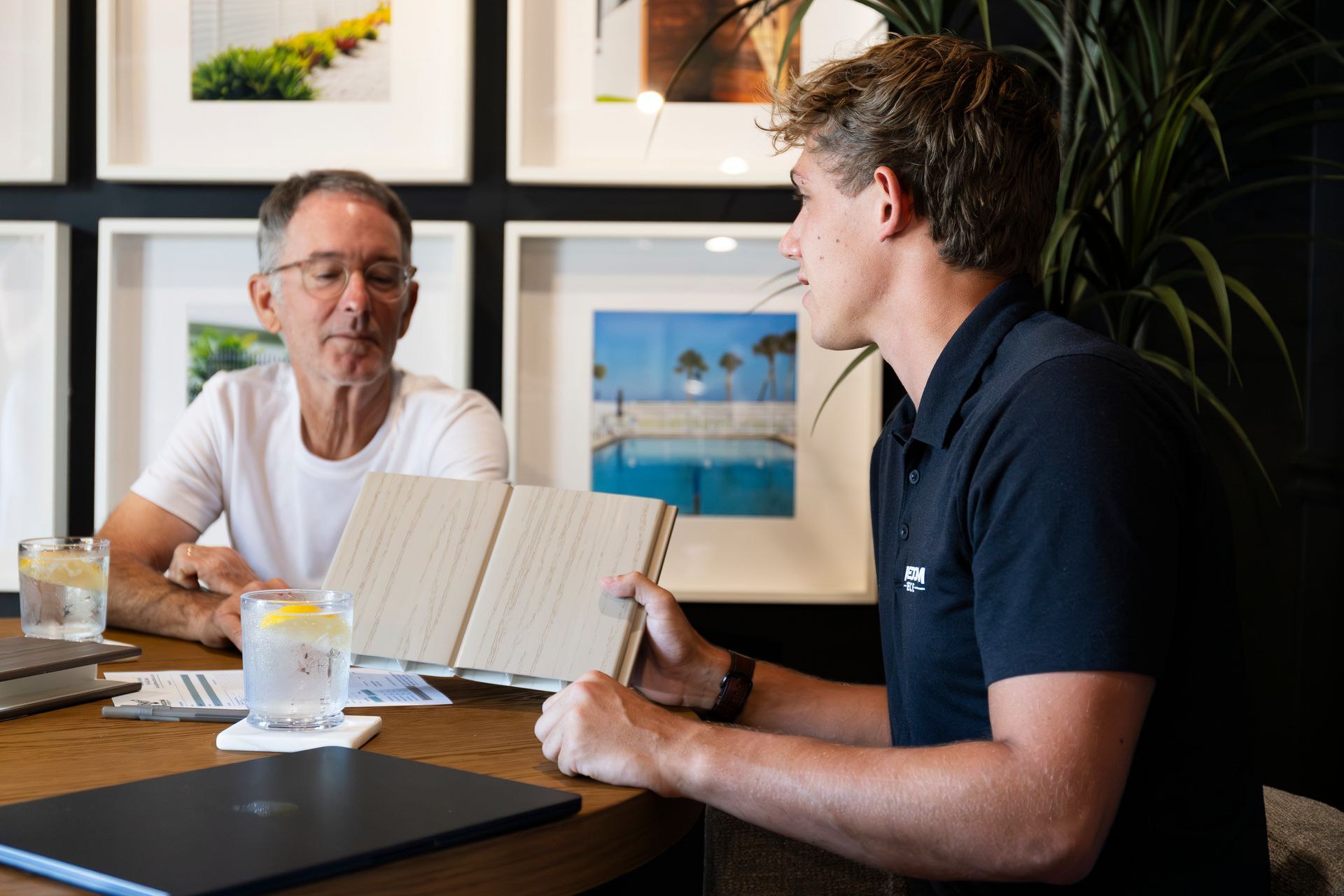 Two people seated at a wooden table in an office setting, looking at an open notebook while discussing a project.
