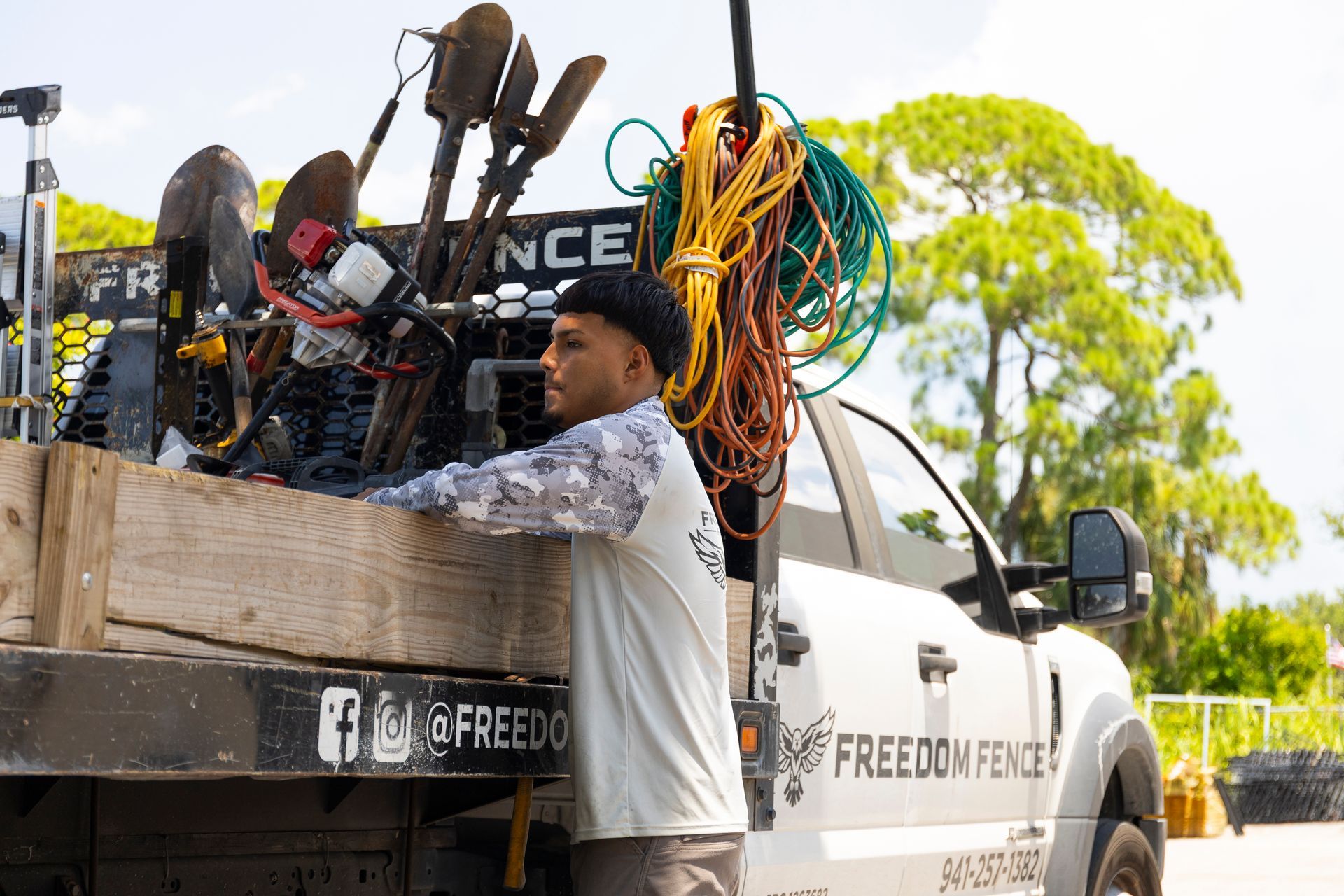 A worker wearing a long-sleeved shirt leans against the bed of a Freedom Fence utility truck loaded with tools and rope.