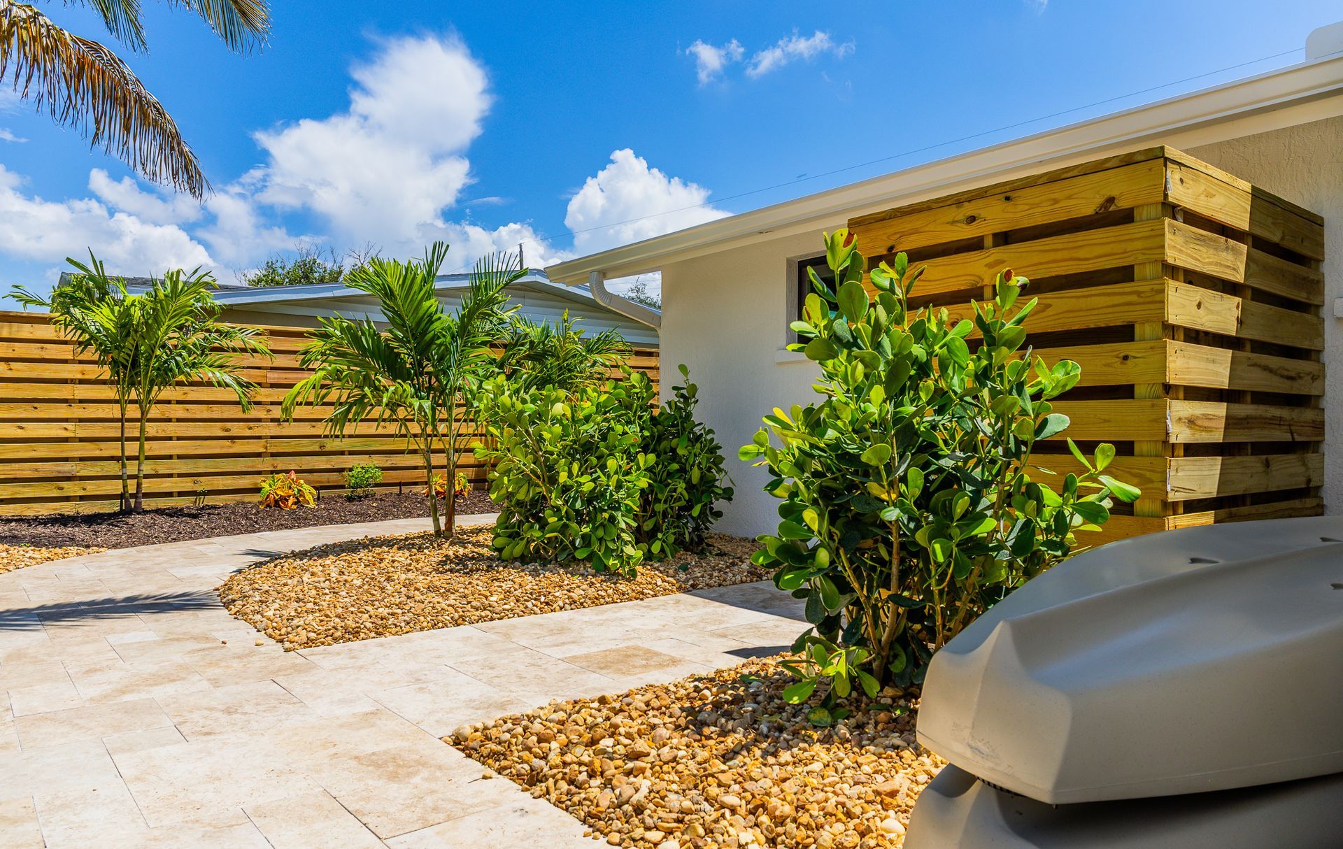A sunny outdoor patio area featuring light stone pavers, golden gravel beds, lush green shrubs, and a wood-slat wall.