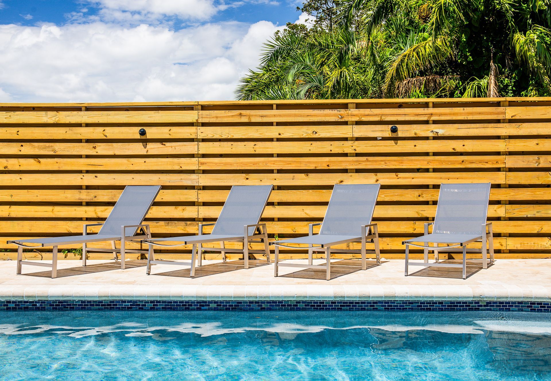 Four grey lounge chairs lined up on a light-colored pool deck in front of a horizontal wooden fence.